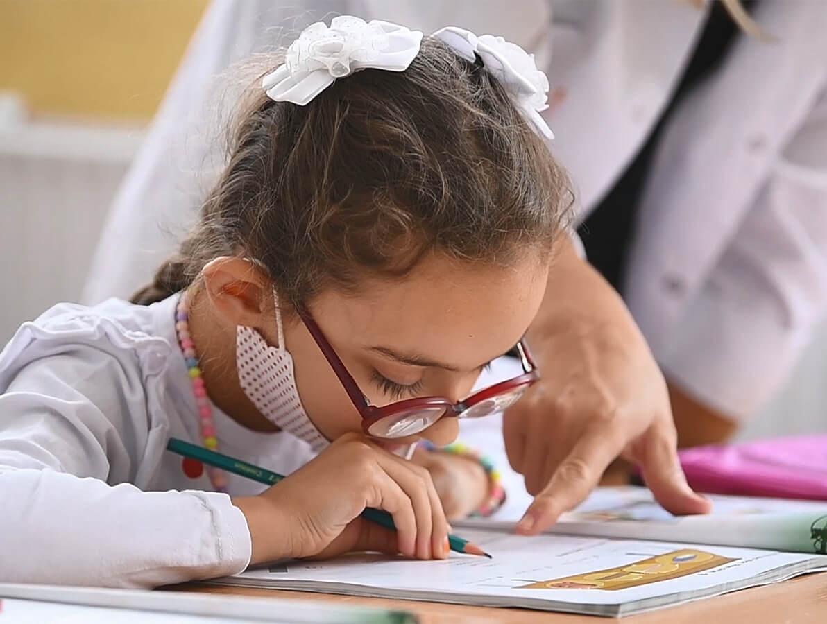 Lapsi, jolla on punaiset silmälasit, katsoo kirjoituskirjaan pitäen kynää kädessään.