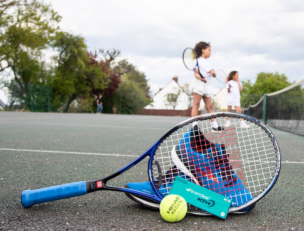 Tennisrack, tennisboll och kontaktlinsförpackning av everclear active med tjejer som spelar tennis i bakgrunden