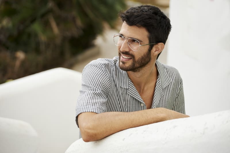 A man in C. Jacobsen aviator eyeglasses, smiling and leaning against a white wall. His relaxed style and sleek frames reflect effortless Scandinavian design.