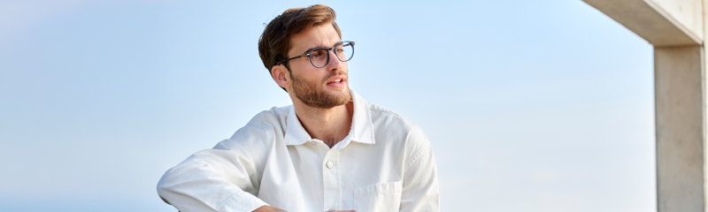 A man wearing round C. Jacobsen eyeglasses with a blue acetate frame, sitting outdoors near the beach. He is dressed in a relaxed white button-up shirt and dark pants.