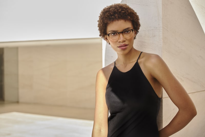 A woman wearing round C. Jacobsen eyeglasses with a subtle keyhole bridge, leaning against a modern architectural wall. She is dressed in a sleek black halter dress, exuding confidence and elegance in a sunlit setting that highlights the frame’s refined.