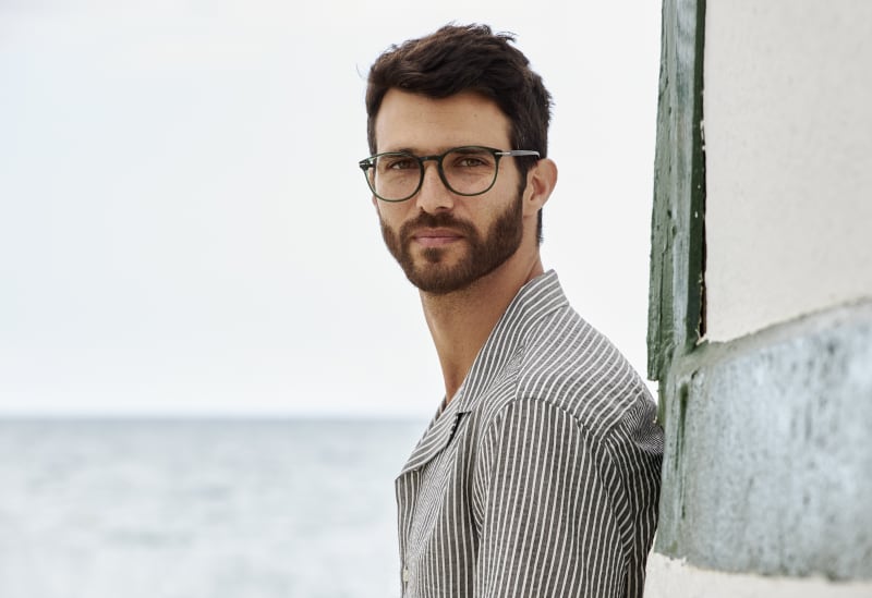 A man wearing Norrsken CJ9012 eyeglasses in a deep green frame, standing near the sea with a calm and confident gaze. The rich color and refined shape of the frame add a modern edge to his relaxed yet sophisticated look.