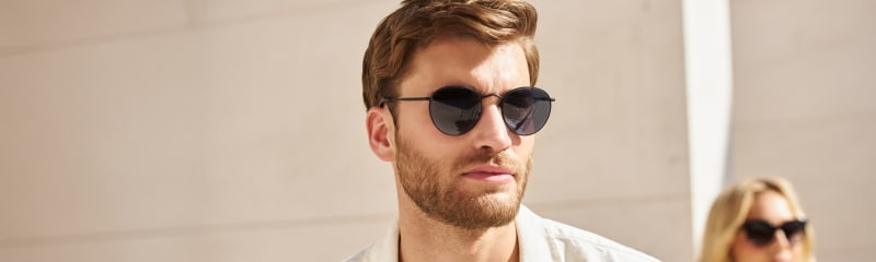 A man wearing round black sunglasses with a thin metal frame, paired with a white jacket and a yellow T-shirt.