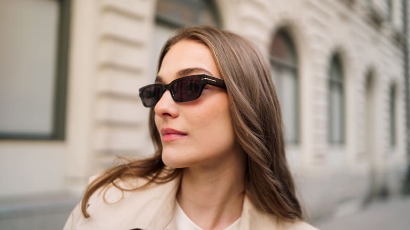 A woman with long brown hair wearing rectangular black sunglasses with a bold frame, walking in an urban setting. She is dressed in a beige trench coat and white top, with a confident expression.