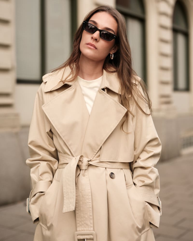 A stylish woman in a beige trench coat and white top, wearing sleek black rectangular sunglasses. She stands against a backdrop of classic city architecture, exuding effortless elegance.
