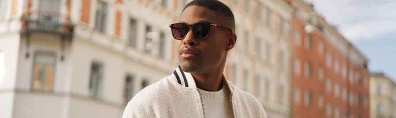 A man wearing brown sunglasses with a classic shape, paired with a textured off-white bomber jacket and a cream sweater.
