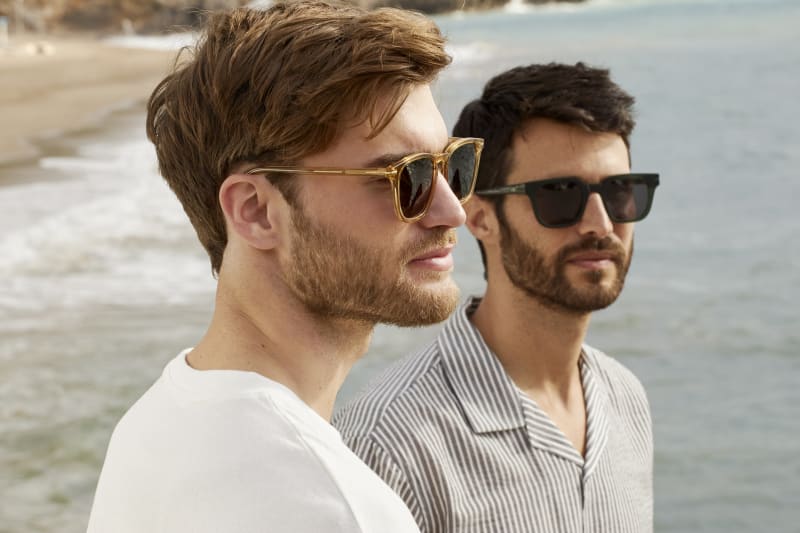 Two men standing by the beach, both wearing C. Jacobsen sunglasses. The man in the foreground wears a thin yellow square-shaped frame, paired with a white T-shirt, while the man in the background wears a classic black square/wayfarer-shaped frame with a striped shirt.