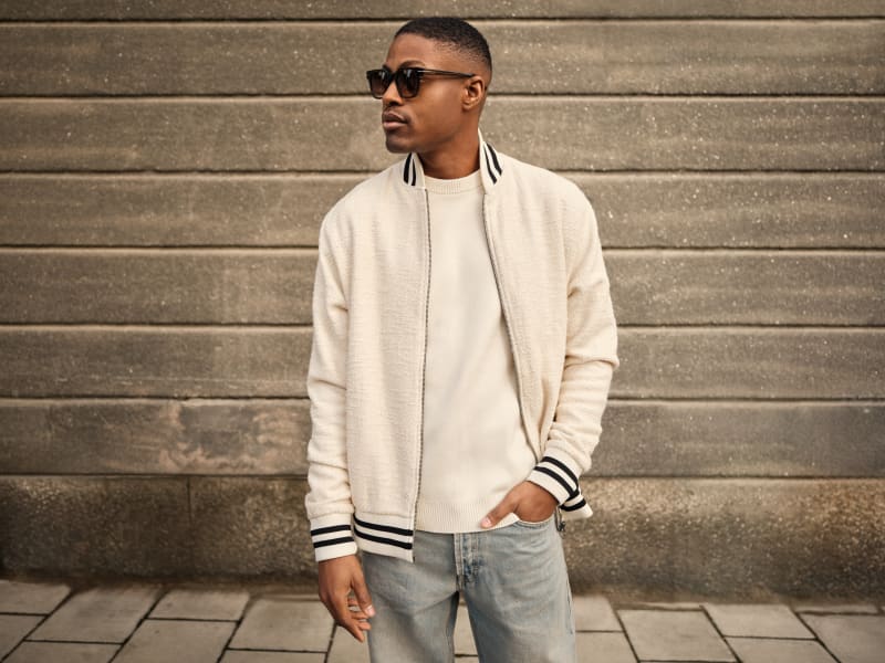 A man is wearing Atrium CJ2409 sunglasses with a classic shape and structured details. He is dressed in a cream bomber jacket, a knit sweater, and light-wash jeans.