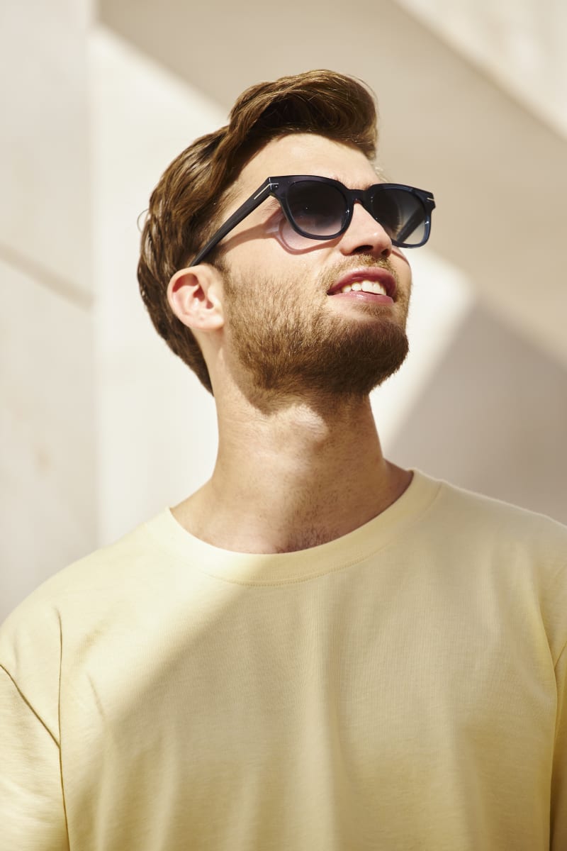 A man wearing dark blue sunglasses with a bold, modern design looks up towards the sunlight. He is dressed in a pale yellow t-shirt, with a relaxed expression.