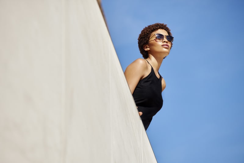 A woman in a black halter dress wears sleek, modern sunglasses with a softly curved frame while looking up against a clear blue sky.