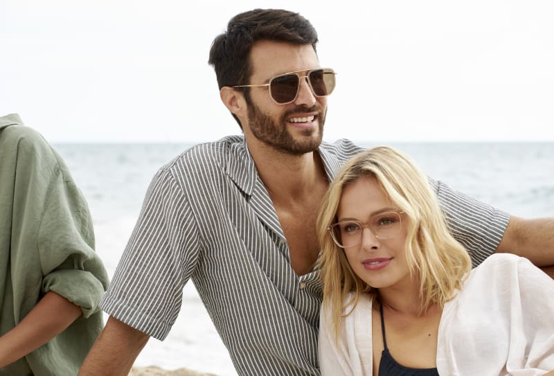 A man wearing Eklips CJ2416 aviator sunglasses with a sleek metal frame, sitting on a beach and smiling. He is dressed in a striped button-down shirt, leaning casually with the ocean in the background. A woman sits beside him wearing eyeglasses, adding to the relaxed and stylish atmosphere.