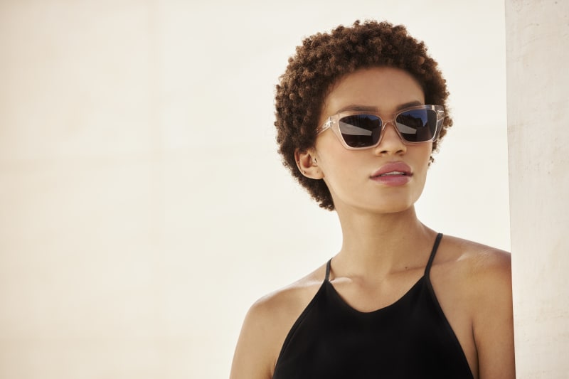 A woman with short curly hair wears Horisont CJ2404 sunglasses, leaning against a minimalist concrete wall. The structured frame complements her modern black halter top.