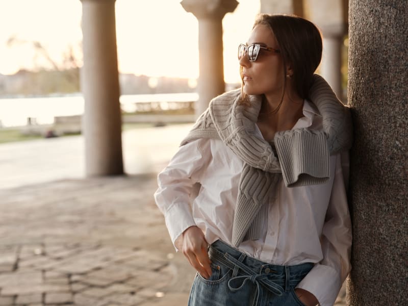 A woman wearing Horisont CJ2404 sunglasses leans against a stone pillar in the warm glow of the setting sun. She is dressed in a white button-up shirt with a knit sweater draped over her shoulders and relaxed-fit denim jeans. The classic yet modern sunglasses complement her effortlessly elegant style, blending timeless sophistication with a contemporary edge.
