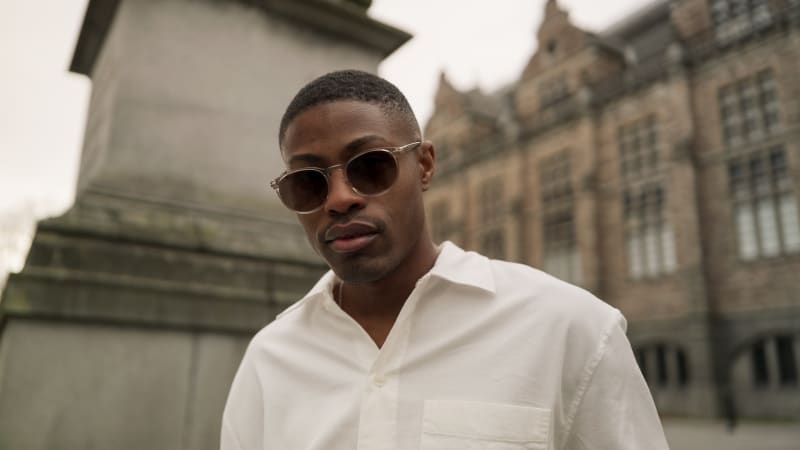 A man wearing Mareld CJ2411 sunglasses, dressed in a white button-up shirt, standing against an urban backdrop with a historic building. His expression is confident as he gazes into the distance.