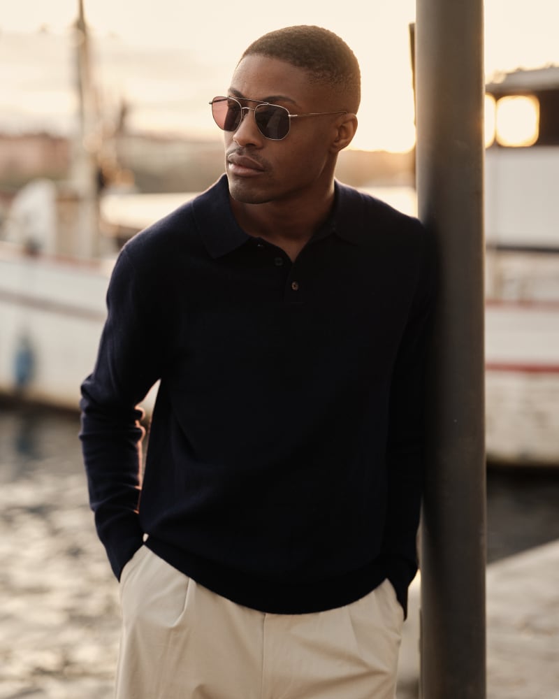 A man in a dark sweater and light trousers leans against a pole by the waterfront, wearing the Porla CJ2417 sunglasses, with the sun setting behind him.