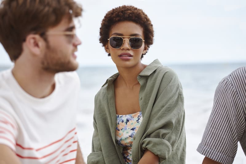 A woman in an olive green shirt and floral top sits on the beach, wearing the Porla CJ2417 sunglasses, with two blurred figures in the foreground.