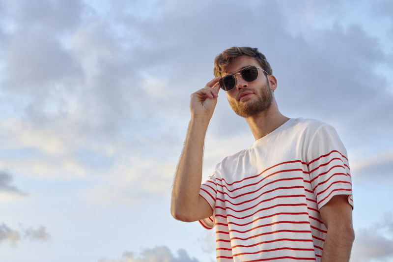 A man in a striped t-shirt adjusts his Porla CJ2417 sunglasses against a blue sky, bathed in golden sunlight.