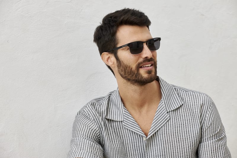 A man with dark hair and a beard wearing Skugga CJ2414 sunglasses and a striped button-up shirt, looking off to the side against a neutral background.