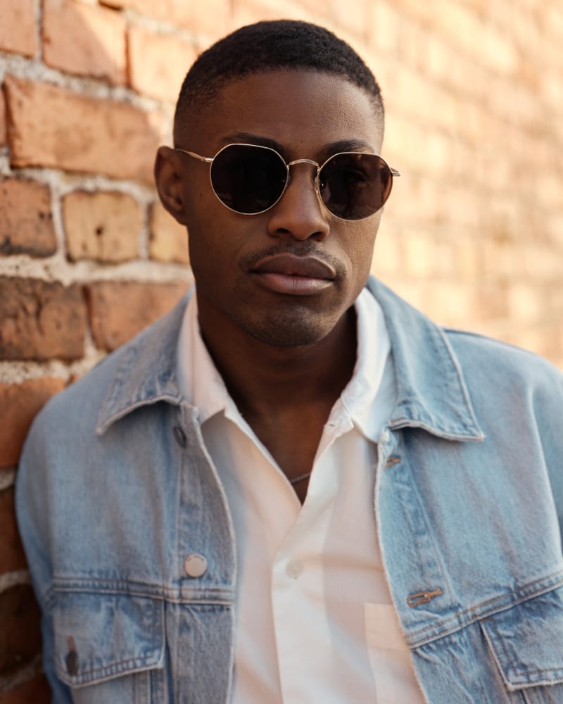 Close-up portrait of a man in Nebula CJ2421 sunglasses, showcasing the frame's round shape and fine metal details against a textured brick background.