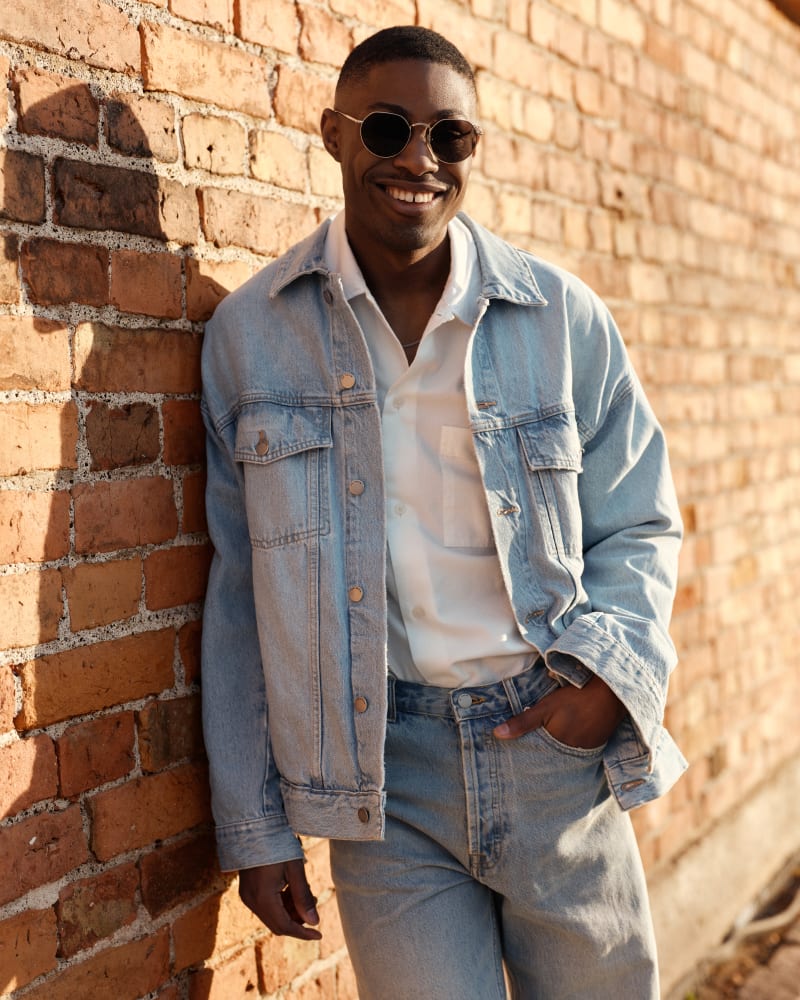 A man wearing Nebula CJ2421 sunglasses leans against a brick wall, dressed in a denim jacket and white shirt, smiling in the warm sunlight.