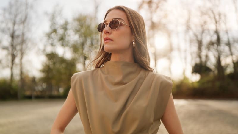 A woman wearing Dagg CJ2428 sunglasses stands in an outdoor setting with trees in the background. The sunlight filters through, enhancing the serene and elegant look of her minimalistic outfit.