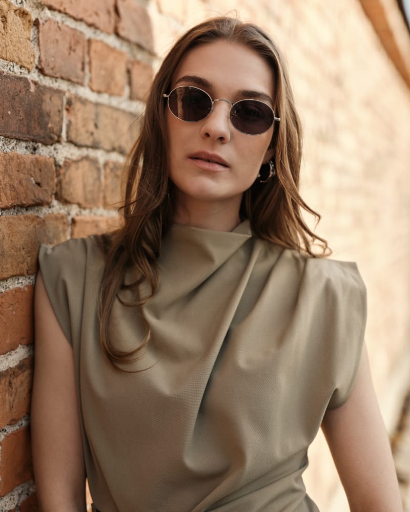 A woman with long, wavy brown hair wearing Dagg CJ2428 sunglasses, leaning against a rustic brick wall. She gazes confidently into the camera, dressed in a draped olive-green top.