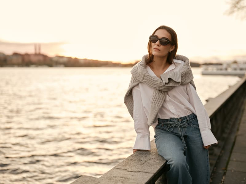 A stylish woman in Vind CJ2431 sunglasses leaning on a waterfront railing, dressed in a white shirt and jeans, with the golden evening light in the background.