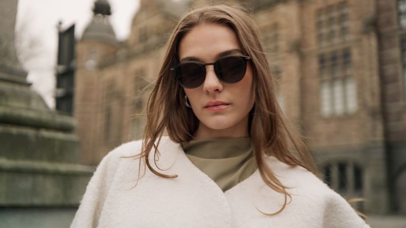 Close-up of a woman wearing Himlakropp CJ2435 sunglasses, showcasing their softly rounded silhouette and minimalistic details. The sophisticated urban setting complements the modern yet classic design of the frame