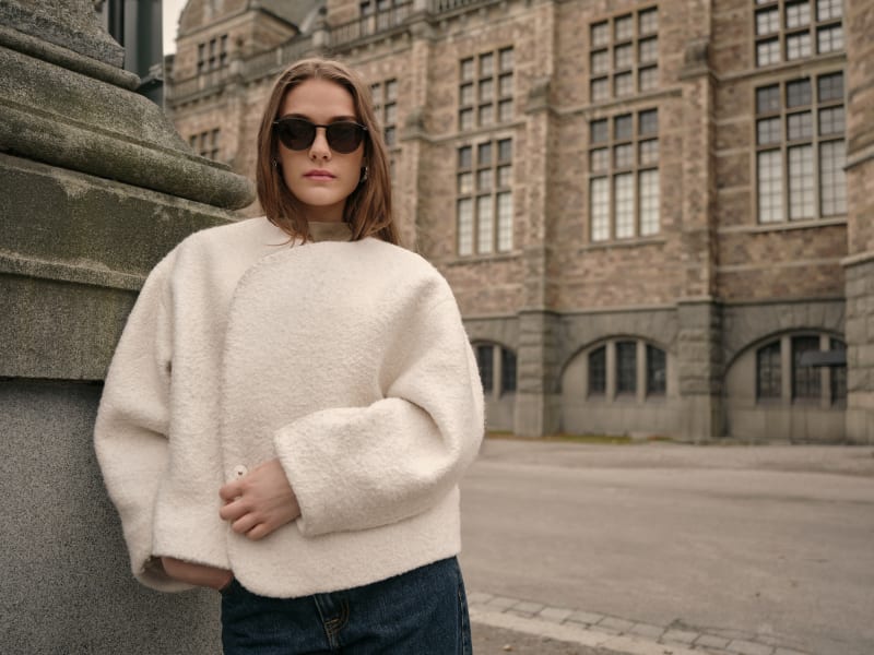 A woman wearing Himlakropp CJ2435 sunglasses and a white wool coat, standing against an elegant stone backdrop. Her neutral-toned outfit enhances the timeless appeal of the round-frame sunglasses.
