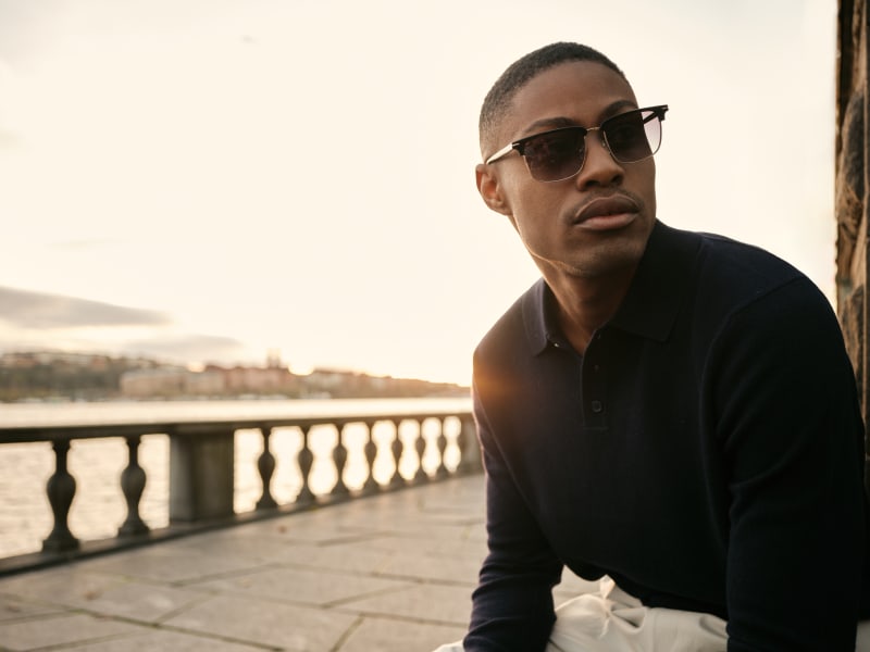 man wearing Clubmaster-inspired sunglasses and a navy polo sits by a waterfront railing at sunset, gazing into the distance.