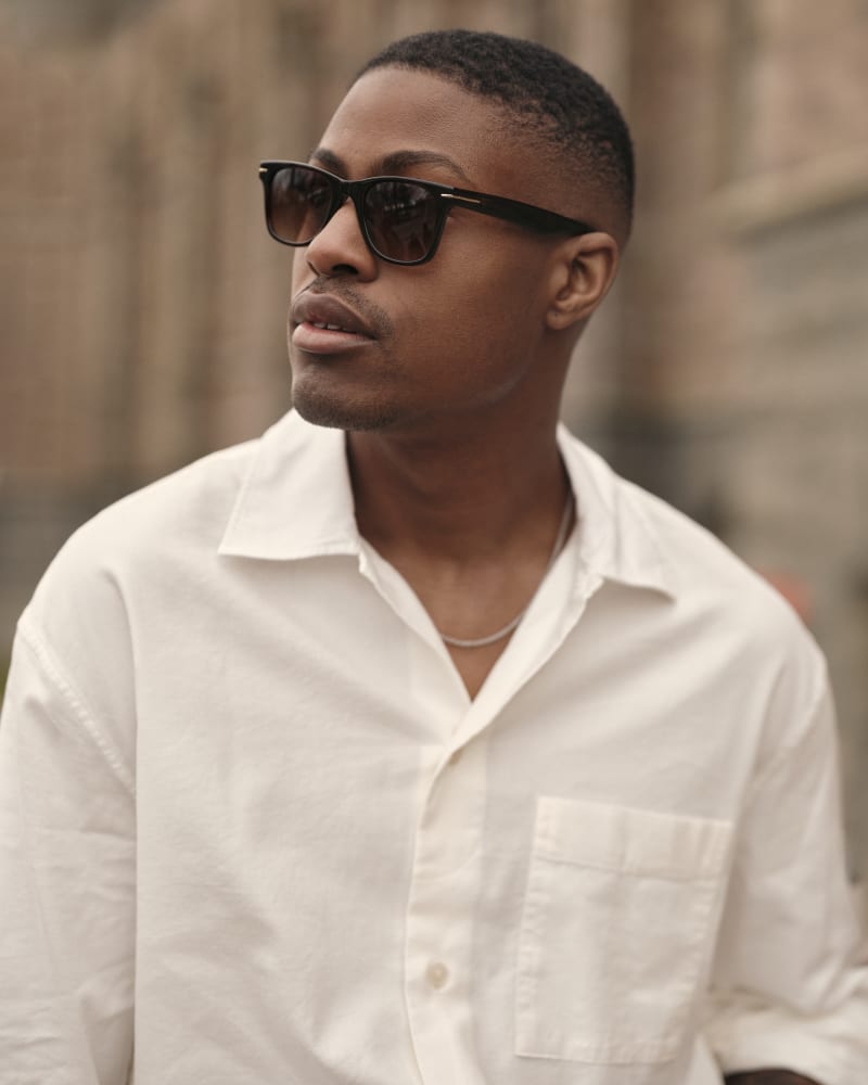 Close-up of a man in Nord CJ2436 sunglasses, wearing a white shirt, with a thoughtful expression against a blurred cityscape background.