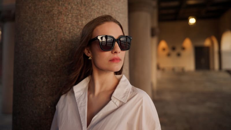 Close-up of a woman wearing the Syd CJ2437 sunglasses, featuring an oversized square frame. She leans against a stone column, gazing into the distance with a confident expression, her white shirt contrasting with the architectural background.