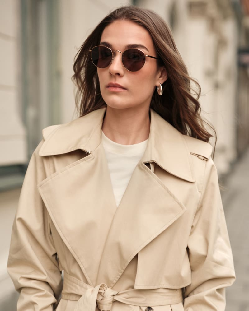 Close-up of a woman wearing Lyster CJ2412 sunglasses with round metal frames. She walks down a city street in a beige trench coat, her hair flowing in the wind, exuding effortless elegance and confidence.