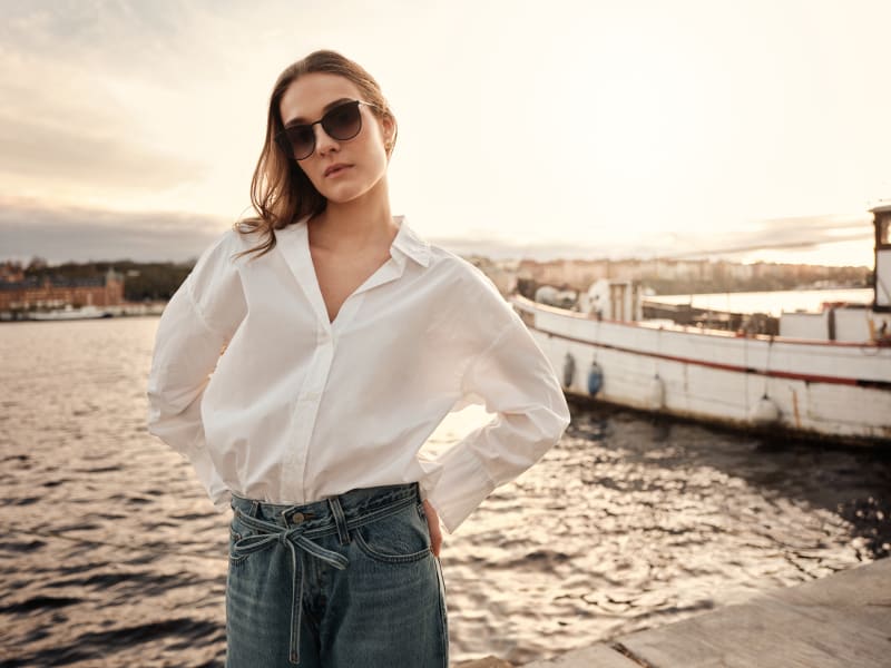A woman wearing Aurora CJ2413 sunglasses stands confidently by the waterfront at sunset. Dressed in a crisp white shirt and relaxed-fit jeans.