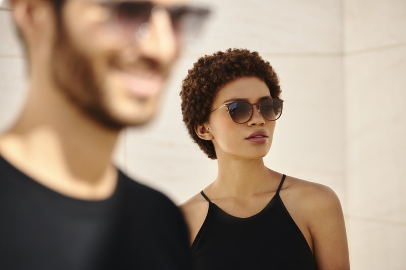 A woman in a black dress wears stylish sunglasses with a rounded, elegant frame while standing in soft sunlight. A blurred man in the foreground, also wearing sunglasses, adds depth to the scene.