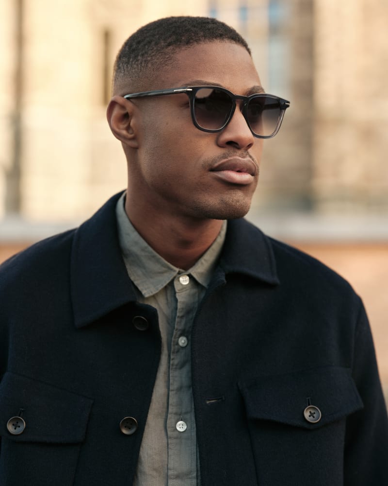 A man in dark blue sunglasses with a squared frame, wearing a dark navy wool coat over a muted green button-up shirt.