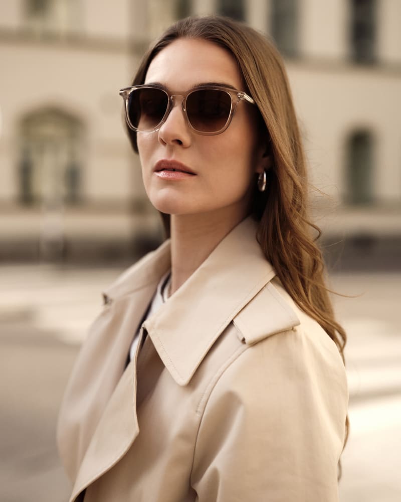 A woman wearing Flack CJ2407 sunglasses and a classic beige trench coat gazes into the distance with confidence. Her soft waves frame her face, and the blurred cityscape in the background adds a timeless, urban elegance to the scene.