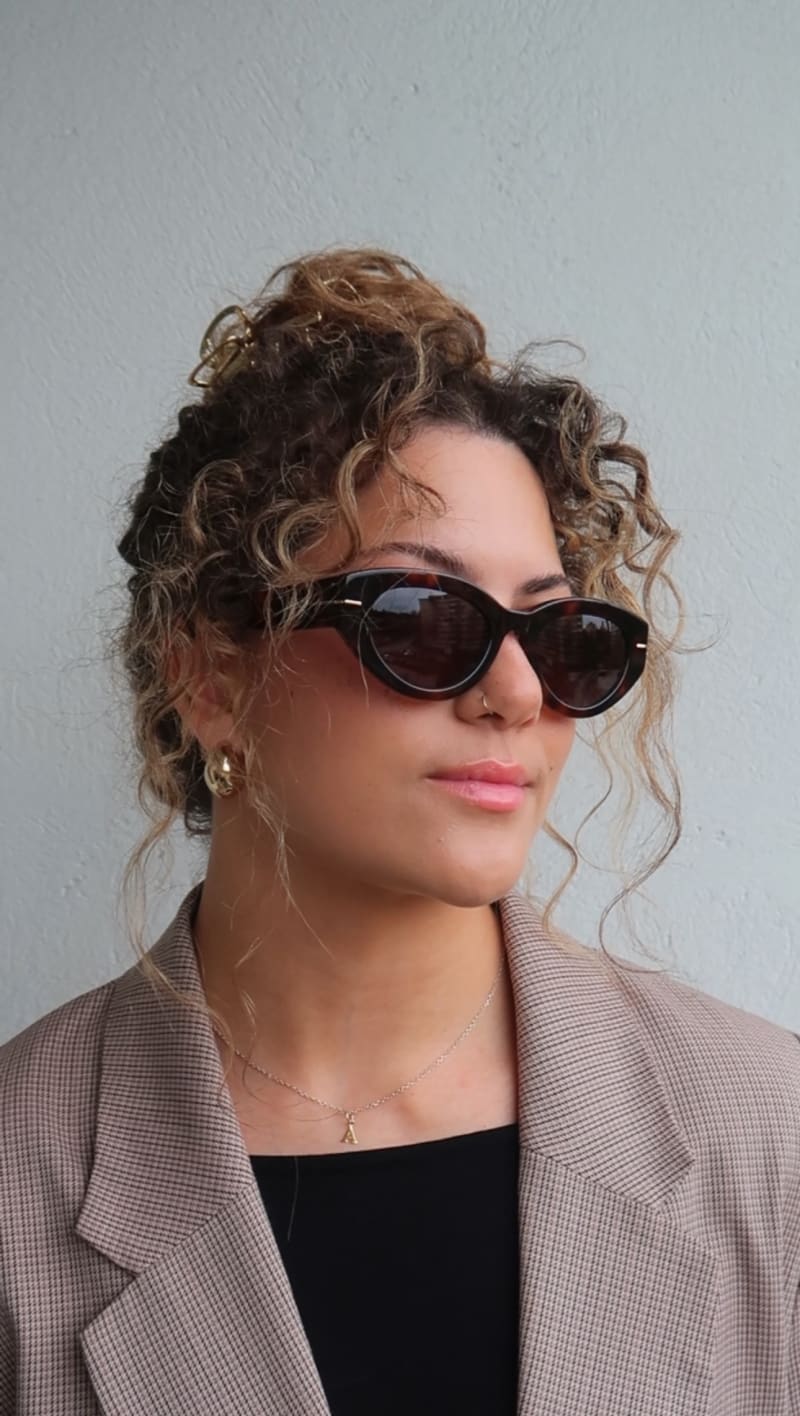 Woman with curly brown hair in an updo, wearing oval black sunglasses, gold hoop earrings, and a beige checked blazer over a black top, looking to the side.