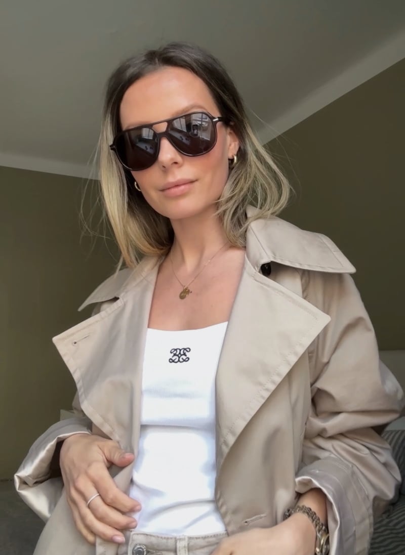 A woman wearing brown sunglasses with a large classic shape, dressed in a white top and a beige jacket.