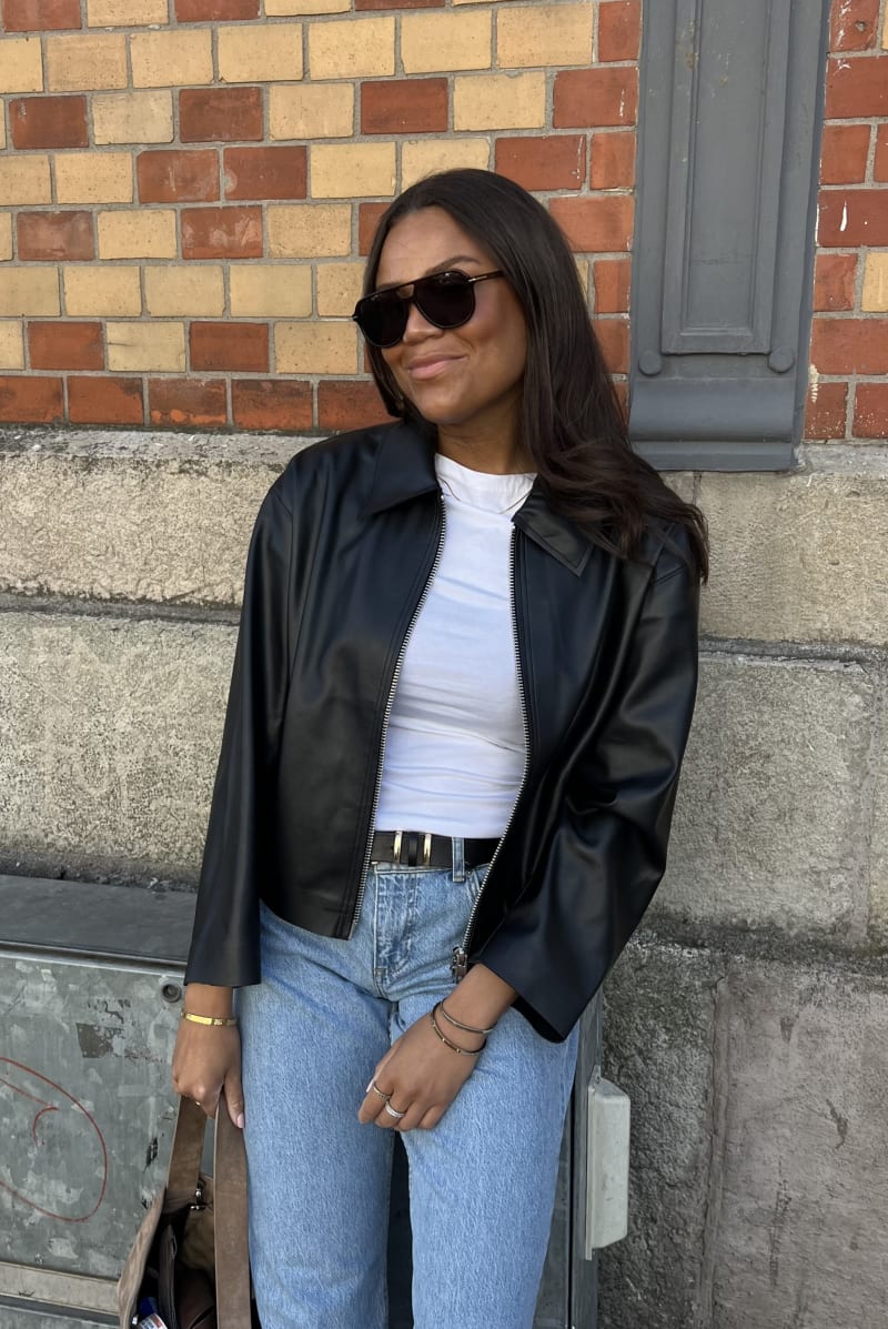 A woman wearing black sunglasses with a large classic shape. She in dressed in a white top, blue jeans and a black leather jacket. She is smiling.