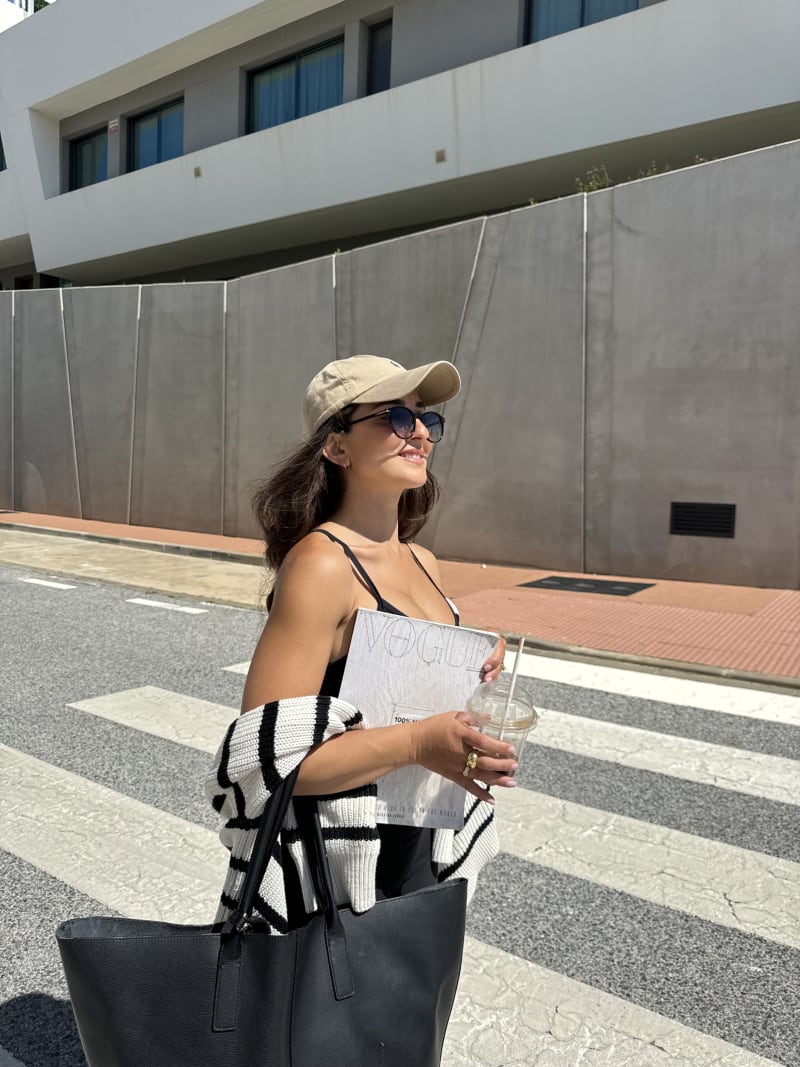 A woman crossing the street and enjoying the sun, wearing her Mareld CJ2411 sunglasses, a beige cap, and a striped cardigan, and carrying a black handbag.