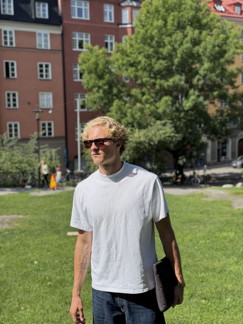 A man with short curly blond hair wearing rectangular brown sunglasses, a plain white T-shirt, and dark jeans, holding a black laptop sleeve while standing on a grassy area in sunlight.