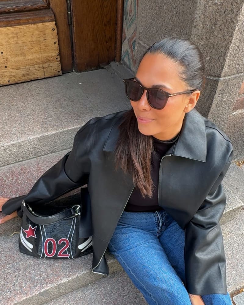 "Woman with long dark hair tied back, wearing rectangular black sunglasses, a black leather jacket, black top, and blue jeans, sitting on stone steps next to a black handbag with red and white star and number 02 design.