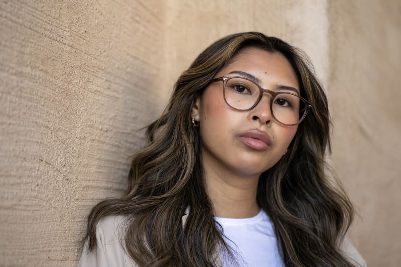 A woman wearing Norrsken CJ9012 eyeglasses, a rounded shaped frame in a light brown color. She is standing in front of a beige wall.