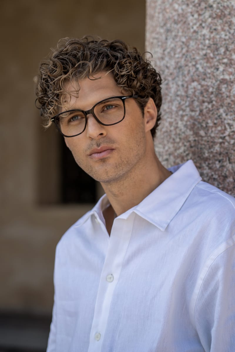 A man, wearing black square-shaped eyeglasses in acetate material. He has curly hair and is dressed in a white shirt.